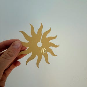 May include: A flat, gold-colored sun-shaped cutout with twelve curved rays radiating from a central circular hole. The object features a small, circular logo with a stylized letter b printed in gold on the lower right side of the sun shape.