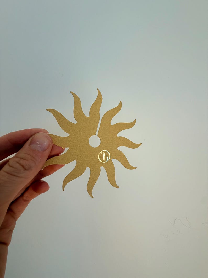 May include: A flat, gold-colored sun-shaped cutout with twelve curved rays radiating from a central circular hole. The object features a small, circular logo with a stylized letter b printed in gold on the lower right side of the sun shape.