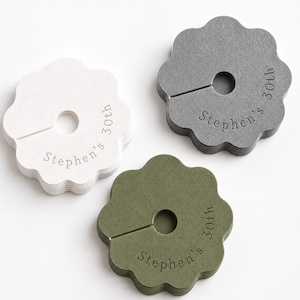 Puede incluir: Tres pilas de etiquetas de papel circulares festoneadas en blanco, gris oscuro y verde oliva. Cada etiqueta tiene un orificio central con una ranura y el texto en relieve "Stephen's 30th" impreso en una fuente serif simple a lo largo del borde.