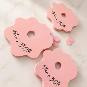 May include: Three stacks of light pink, flower-shaped paper tags with a central circular cutout and a slit. Each tag features the black printed text "Mari's 30th" in a cursive script. Small pink circular paper confetti pieces are scattered nearby.