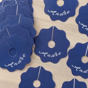 May include: A collection of dark blue paper wine glass markers with a scalloped edge, a circular center cutout, and a vertical slit. Each marker features the name Andre printed in white cursive script. The markers are arranged in a stack and scattered.