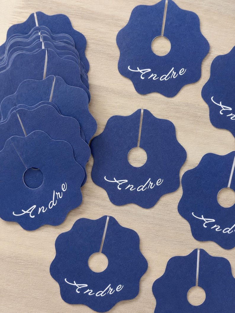 May include: A collection of dark blue paper wine glass markers with a scalloped edge, a circular center cutout, and a vertical slit. Each marker features the name Andre printed in white cursive script. The markers are arranged in a stack and scattered.