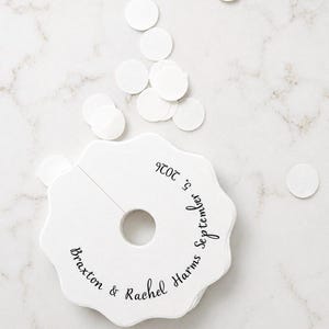 May include: A stack of white, scalloped-edge paper tags with a central circular cutout and a slit. The tags feature black printed text that reads Braxton & Rachel Harms September 5, 2026. Several loose, small white circular paper confetti pieces are nearby.
