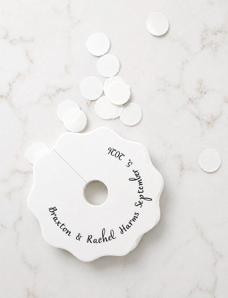 May include: A stack of white, scalloped-edge paper tags with a central circular cutout and a slit. The tags feature black printed text that reads Braxton & Rachel Harms September 5, 2026. Several loose, small white circular paper confetti pieces are nearby.