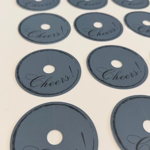 May include: A collection of circular, slate-gray paper tags, each featuring a central hole and a slit extending to the edge. The word Cheers! is printed in a black, elegant script font, accompanied by a thin, hand-drawn style circular border.