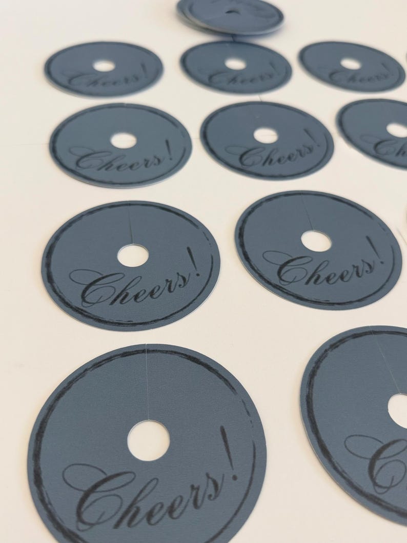 May include: A collection of circular, slate-gray paper tags, each featuring a central hole and a slit extending to the edge. The word Cheers! is printed in a black, elegant script font, accompanied by a thin, hand-drawn style circular border.