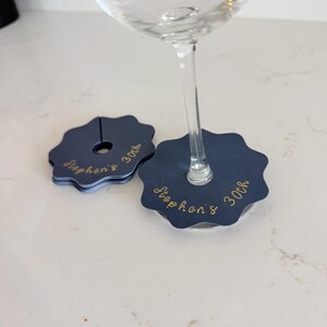 May include: A set of navy blue scalloped wine glass markers with a central slit and hole. Each marker features the gold cursive text "Stephen's 30th" printed on the surface. One marker is attached to the stem of a clear glass, while others are stacked.