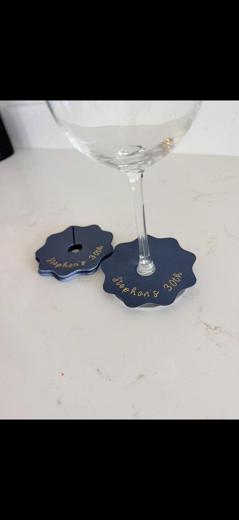 May include: A set of navy blue scalloped wine glass markers with a central slit and hole. Each marker features the gold cursive text "Stephen's 30th" printed on the surface. One marker is attached to the stem of a clear glass, while others are stacked.