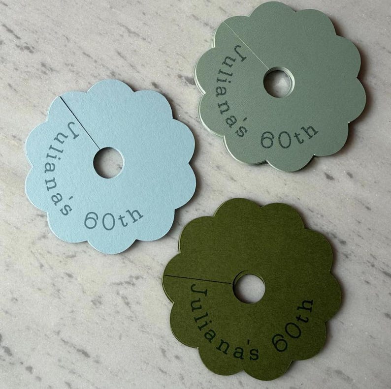 May include: Three scalloped circular paper tags in light blue, sage green, and olive green. Each tag features a central circular cutout, a slit extending to the edge, and the printed text "Juliana's 60th" in a dark, sans-serif font.