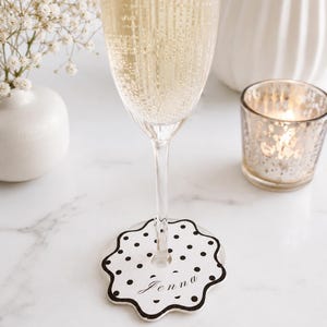 May include: A white circular wine glass marker with a scalloped edge and a black polka dot pattern. The marker features the name Jenna written in a black cursive script font in the center and includes a central hole for the glass stem.