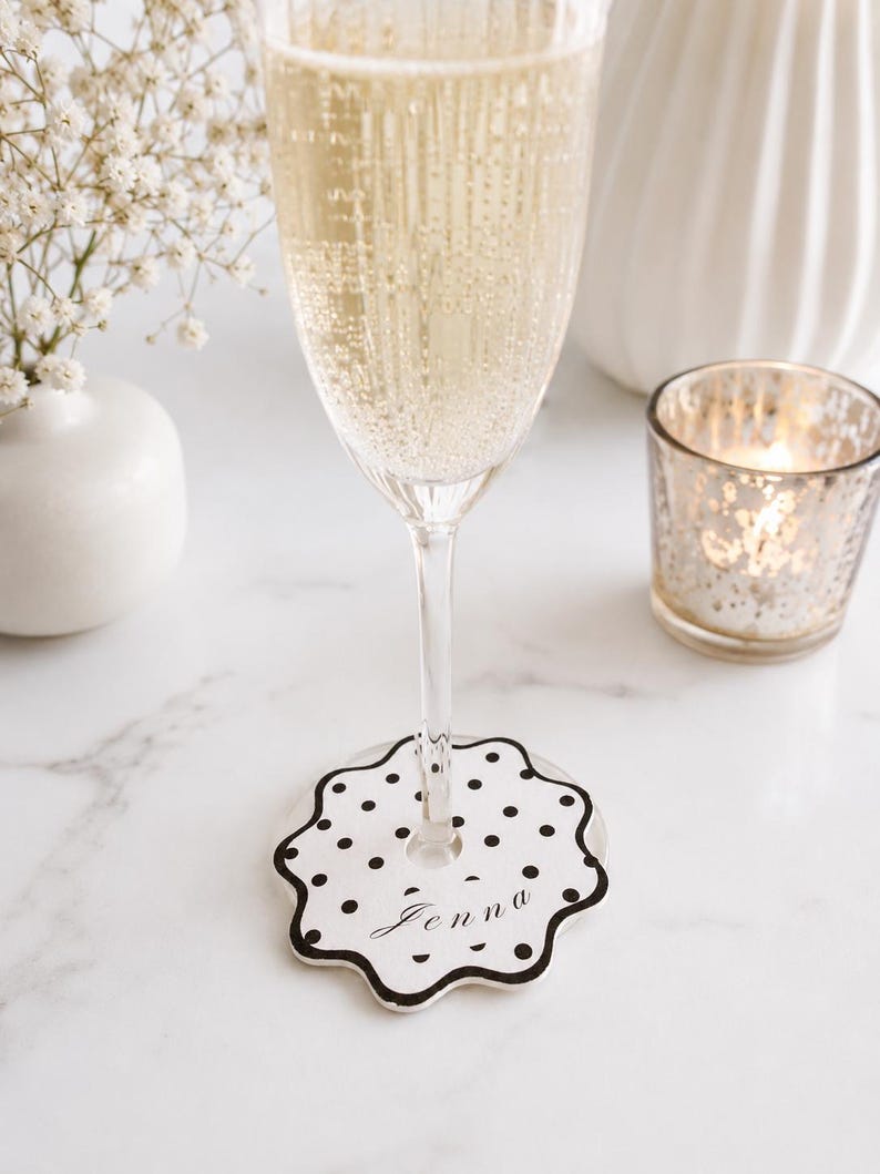 May include: A white circular wine glass marker with a scalloped edge and a black polka dot pattern. The marker features the name Jenna written in a black cursive script font in the center and includes a central hole for the glass stem.