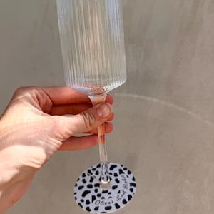 May include: A clear, ribbed glass champagne flute featuring a slender stem and a circular base decorated with a black and white cow print pattern. The base includes the printed text "Olivia's Last Rodeo" in a black cursive script font.