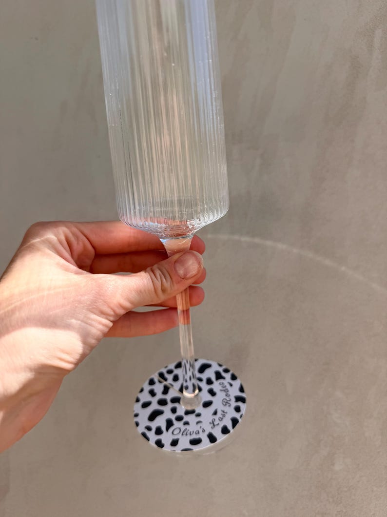 May include: A clear, ribbed glass champagne flute featuring a slender stem and a circular base decorated with a black and white cow print pattern. The base includes the printed text "Olivia's Last Rodeo" in a black cursive script font.