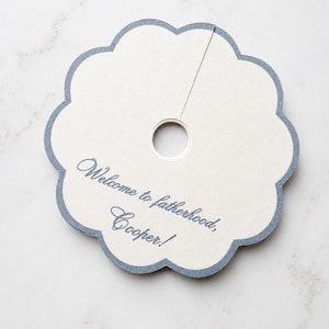 May include: A circular, scalloped-edge paper tag with a central hole and a slit extending to the edge. The tag is off-white with a blue border and features the blue printed text: Welcome to fatherhood, Cooper!