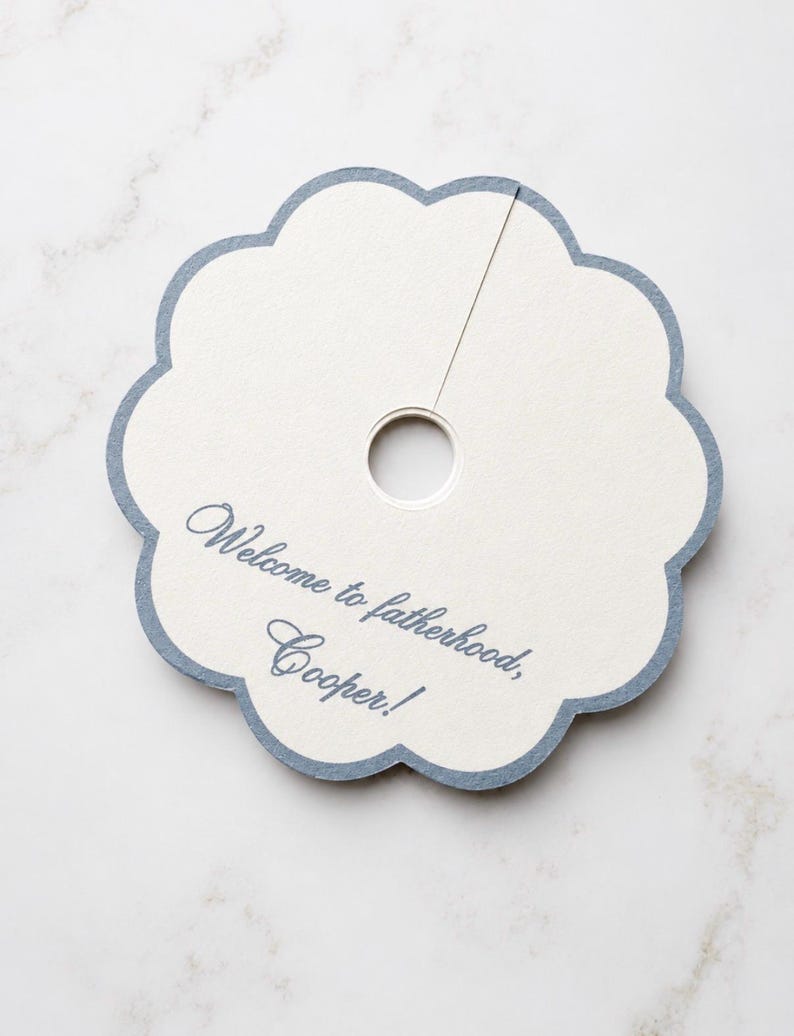 May include: A circular, scalloped-edge paper tag with a central hole and a slit extending to the edge. The tag is off-white with a blue border and features the blue printed text: Welcome to fatherhood, Cooper!