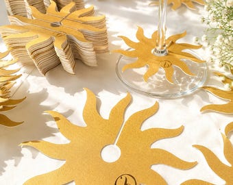 May include: A collection of gold-colored sun-shaped wine glass markers made of cardstock. Each marker features a central circular cutout with a slit for attachment to a glass stem and a small black logo printed on one of the sun's rays.
