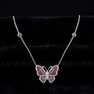 May include: A silver-toned necklace featuring a central butterfly pendant encrusted with small pink stones and a border of clear stones. The chain includes two bezel-set pink stones on each side, with the text JEWELICIOUS STORE visible in the background.