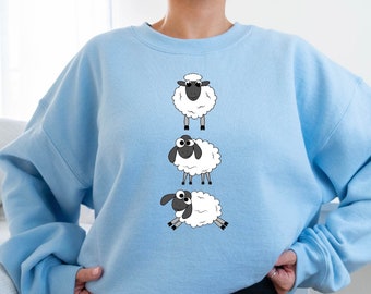 Könnte beinhalten: Ein hellblaues Sweatshirt mit einem Motiv aus drei Cartoon-Schafen. Die Schafe sind weiß mit schwarzen Gesichtern und Beinen. Die Schafe stehen in einer Reihe, wobei das oberste Schaf aufrecht steht, das mittlere Schaf zur Seite schaut und das unterste Schaf liegt.