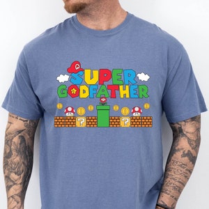 Puede incluir: Una camiseta azul con un gr&aacute;fico de un personaje de Super Mario Bros. y el texto "SUPER GODFATHER".