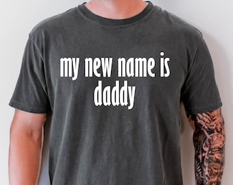 May include: A dark gray t-shirt with the text "my new name is daddy" printed in white.