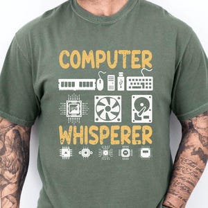 May include: Olive green t-shirt with the text "COMPUTER WHISPERER" in yellow and white. The design includes computer-related icons such as a keyboard, mouse, and fan. The shirt is a casual wear item.
