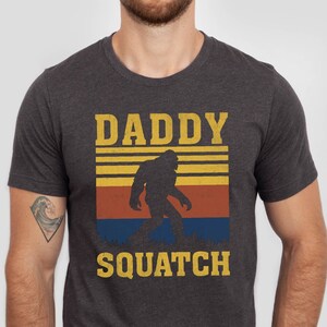 May include: A dark heather grey t-shirt with a retro-style graphic of a Bigfoot walking in front of a sunset. The text "Daddy Squatch" is printed on the shirt.