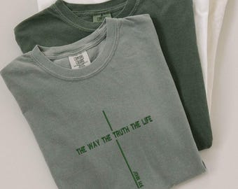 May include: Four folded t-shirts in cream, white, and green hues. The front shirt is a muted green with the text "THE WAY THE TRUTH THE LIFE" and "JOHN 14:6" in green. Each shirt has a tag that reads "COMFORT COLORS".