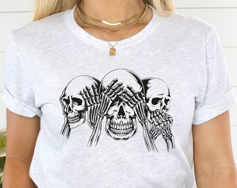 May include: A white t-shirt with a black graphic of three skulls, each with a hand covering their eyes, nose, and mouth. The graphic is a play on the "See no evil, hear no evil, speak no evil" saying.