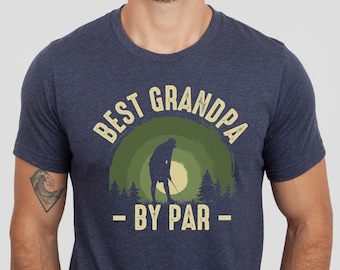 May include: A navy blue t-shirt with a vintage-style graphic of a golfer in front of a green and yellow sunset. The text "BEST GRANDPA - BY PAR -" is printed on the shirt.