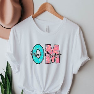 May include: White t-shirt with a graphic design of the letters "OM" in turquoise and pink with a pattern. The letters stand for "Office Manager".