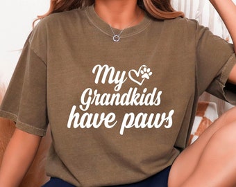 May include: A woman wearing a brown t-shirt with the text "My grandkids have paws" in white. The shirt has a heart with a paw print inside the heart. She is wearing blue shorts and white trainers.