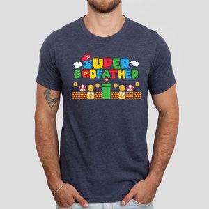 Puede incluir: Una camiseta azul marino con un gr&aacute;fico de un personaje de videojuego. El gr&aacute;fico dice "SUPER GODFATHER" y presenta una gorra roja, una estrella amarilla y una tuber&iacute;a verde.