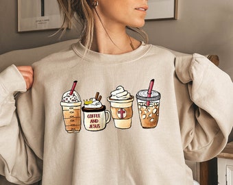 May include: A beige sweatshirt featuring a graphic of four coffee drinks. The drinks are a caramel macchiato, a hot chocolate with whipped cream, a coffee with a cross on it, and an iced coffee. The text "Coffee and Jesus" is printed on the sweatshirt.