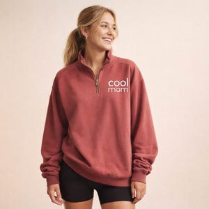 May include: A brick-red sweatshirt with a quarter-zip neckline and the words "cool mom" printed in white. The long-sleeved top is made of a soft material.