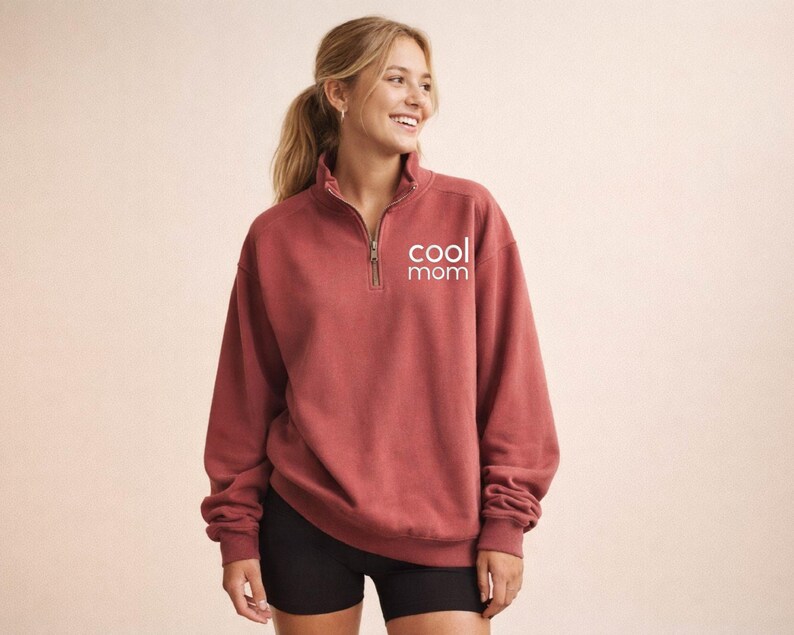 May include: A brick-red sweatshirt with a quarter-zip neckline and the words "cool mom" printed in white. The long-sleeved top is made of a soft material.