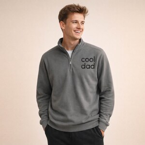 May include: A gray pullover sweatshirt with a quarter-zip collar and the text "cool dad" printed on the chest. The sweatshirt is a solid color and has long sleeves. The person is wearing a white undershirt.