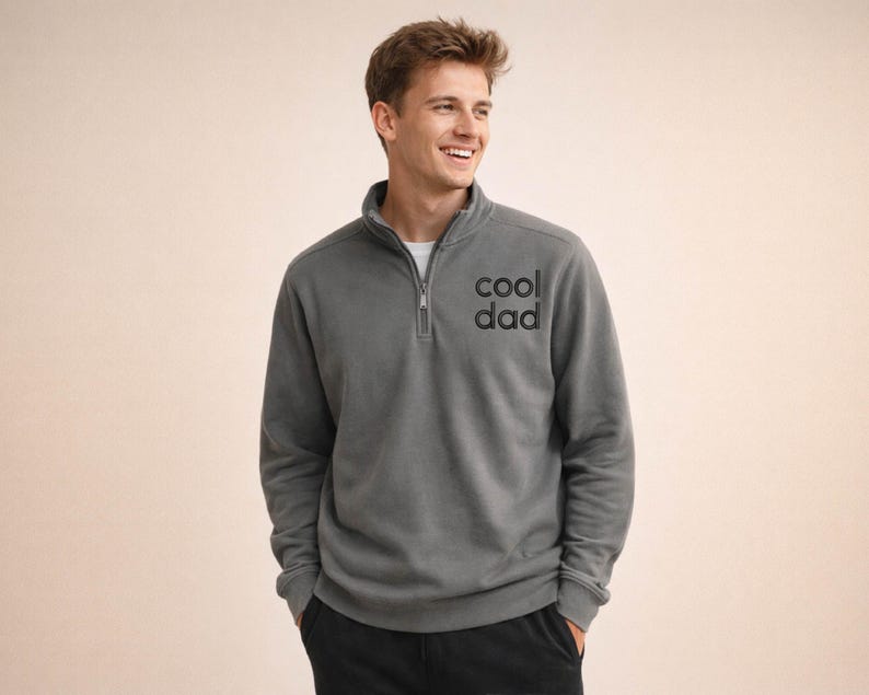 May include: A gray pullover sweatshirt with a quarter-zip collar and the text "cool dad" printed on the chest. The sweatshirt is a solid color and has long sleeves. The person is wearing a white undershirt.