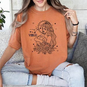 Peut inclure: Un t-shirt orange br&ucirc;l&eacute; avec un graphique noir et blanc d'une femme avec une couronne de fleurs tenant un globe. Le graphique comprend &eacute;galement le texte "VIRGO" et des &eacute;toiles.