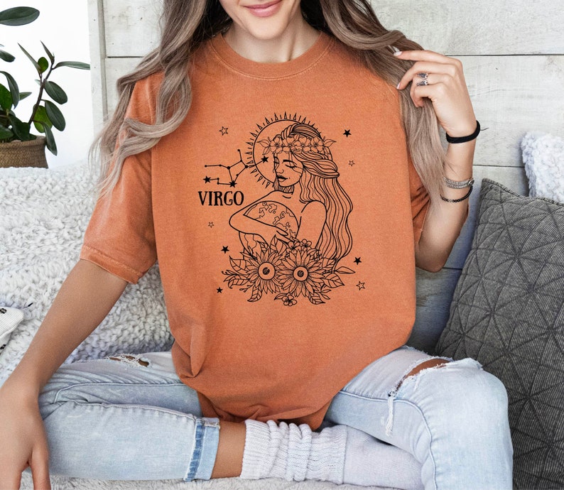 Peut inclure: Un t-shirt orange br&ucirc;l&eacute; avec un graphique noir et blanc d'une femme avec une couronne de fleurs tenant un globe. Le graphique comprend &eacute;galement le texte "VIRGO" et des &eacute;toiles.