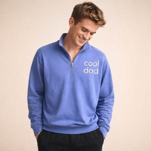 May include: A periwinkle blue pullover sweatshirt with a quarter-zip neckline. The text "cool dad" is printed in white on the chest. The sweatshirt is paired with black pants. The model is smiling.