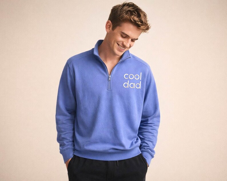 May include: A periwinkle blue pullover sweatshirt with a quarter-zip neckline. The text "cool dad" is printed in white on the chest. The sweatshirt is paired with black pants. The model is smiling.