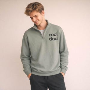 May include: Sage green pullover sweatshirt with a quarter-zip collar and the text "cool dad" printed on the chest. The sweatshirt is a solid color and has long sleeves. The person is wearing black pants.