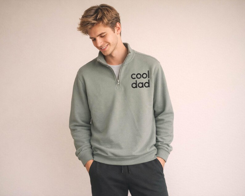 May include: Sage green pullover sweatshirt with a quarter-zip collar and the text "cool dad" printed on the chest. The sweatshirt is a solid color and has long sleeves. The person is wearing black pants.