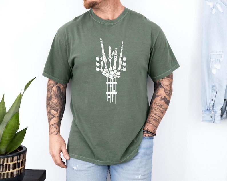Pu&ograve; includere: Una maglietta verde con una grafica bianca di una mano scheletrica che suona la chitarra. La mano sta facendo un segno rock and roll con l'indice e il mignolo estesi.