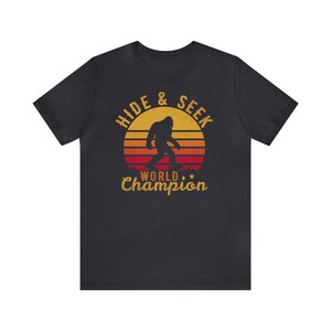 May include: Black t-shirt with a retro-style graphic of a Bigfoot walking in front of a sunset. The graphic says "Hide & Seek World Champion".