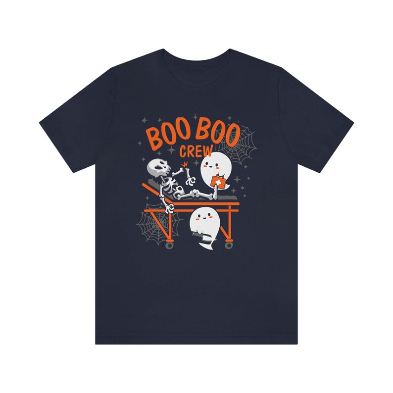 May include: Navy blue t-shirt with a Halloween-themed graphic. The design features a skeleton on a stretcher attended by two cartoon ghosts. The text "BOO BOO CREW" is printed above the characters in orange.
