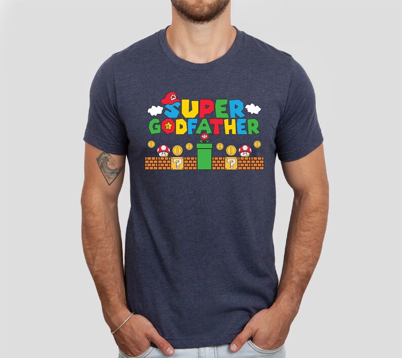 Puede incluir: Una camiseta azul marino con un gr&aacute;fico de un personaje de videojuego. El gr&aacute;fico dice "SUPER GODFATHER" y presenta una gorra roja, una estrella amarilla y una tuber&iacute;a verde.