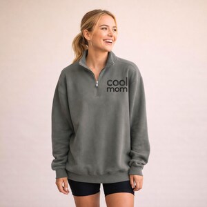 May include: A gray sweatshirt with a quarter-zip neckline and the text "cool mom" in black. The sweatshirt is a relaxed fit and has long sleeves. The model is wearing black shorts.