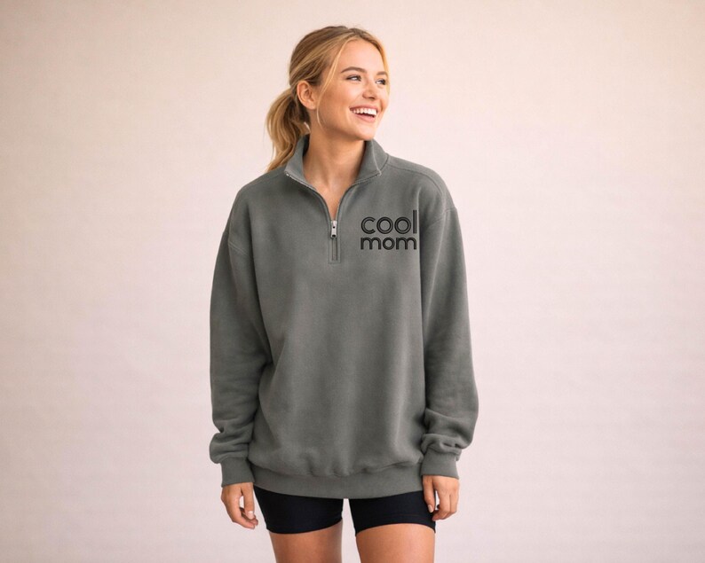 May include: A gray sweatshirt with a quarter-zip neckline and the text "cool mom" in black. The sweatshirt is a relaxed fit and has long sleeves. The model is wearing black shorts.