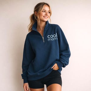 May include: Navy blue quarter-zip sweatshirt with the text "cool mom" in light blue. The sweatshirt has a high collar and a silver zipper. The model is wearing black shorts.